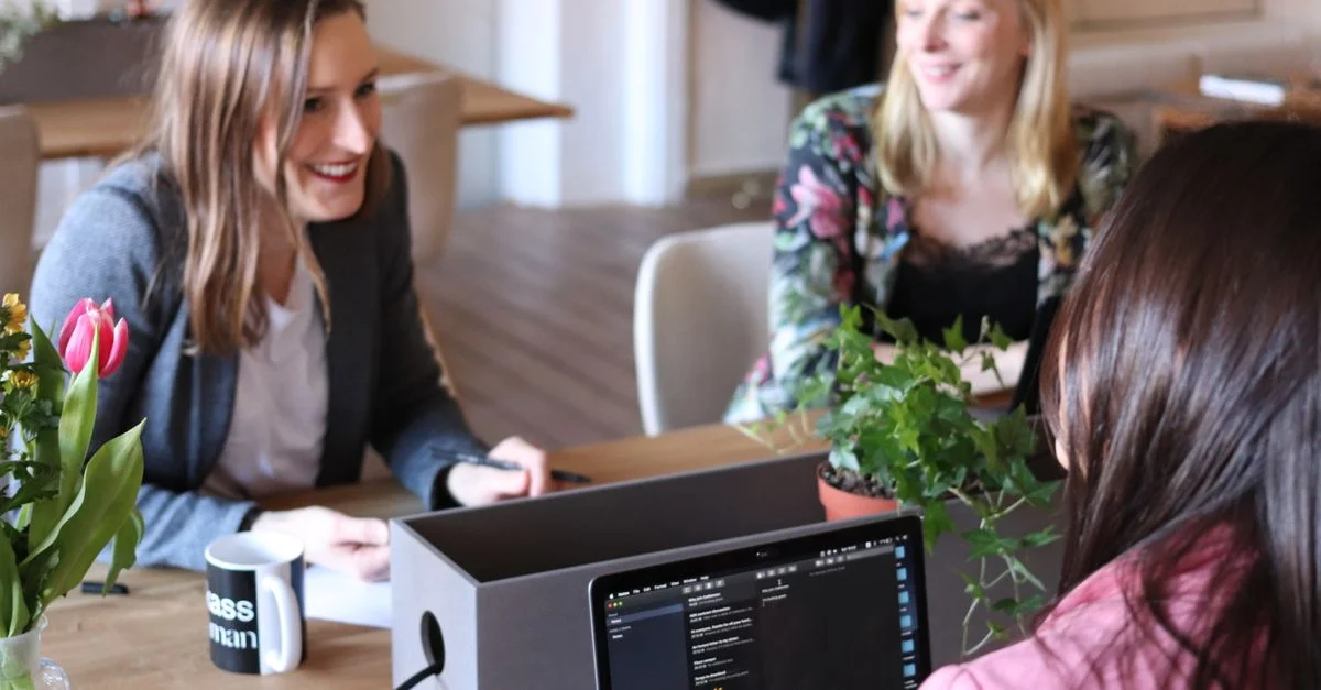 Trois femmes participant à une réunion d'affaires décontractée dans un bureau moderne avec des ordinateurs portables et des plantes.