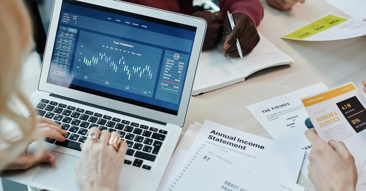 Des professionnels du secteur analysent des données financières sur un ordinateur portable lors d'une réunion. Graphiques et rapports visibles.