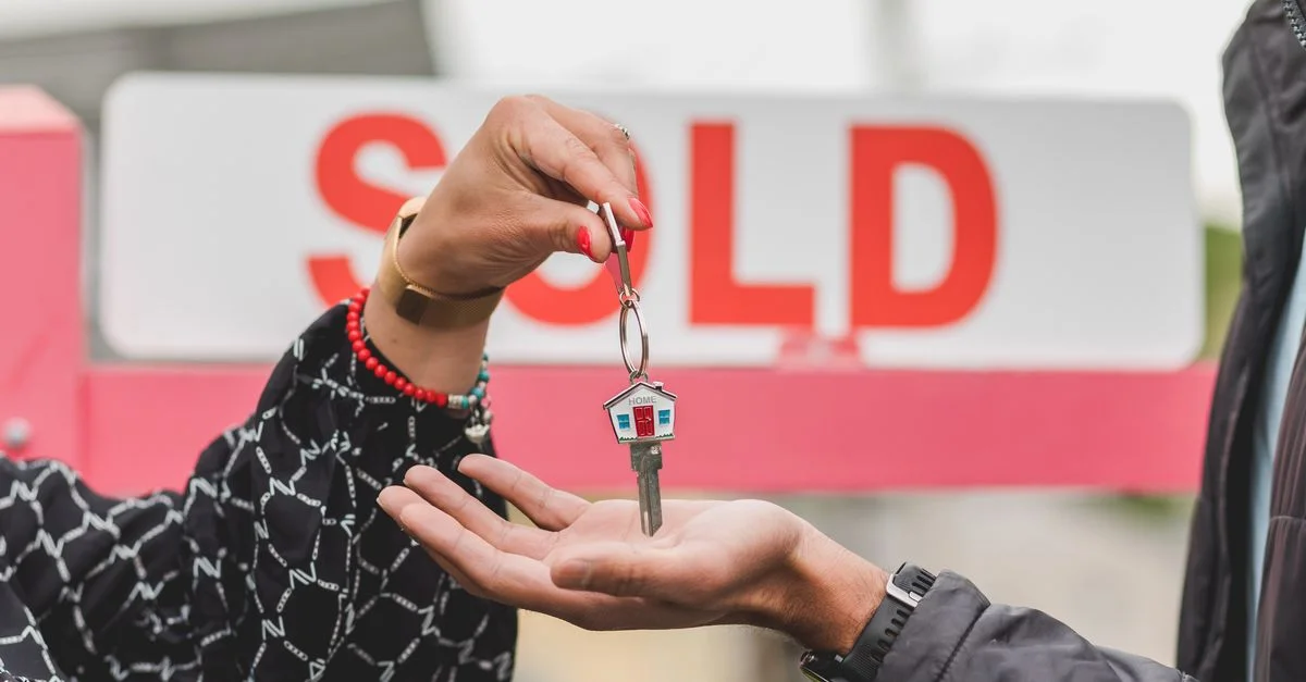 Gros plan sur la remise d'une clé de maison avec un panneau vendu en arrière-plan, symbolisant le succès immobilier.