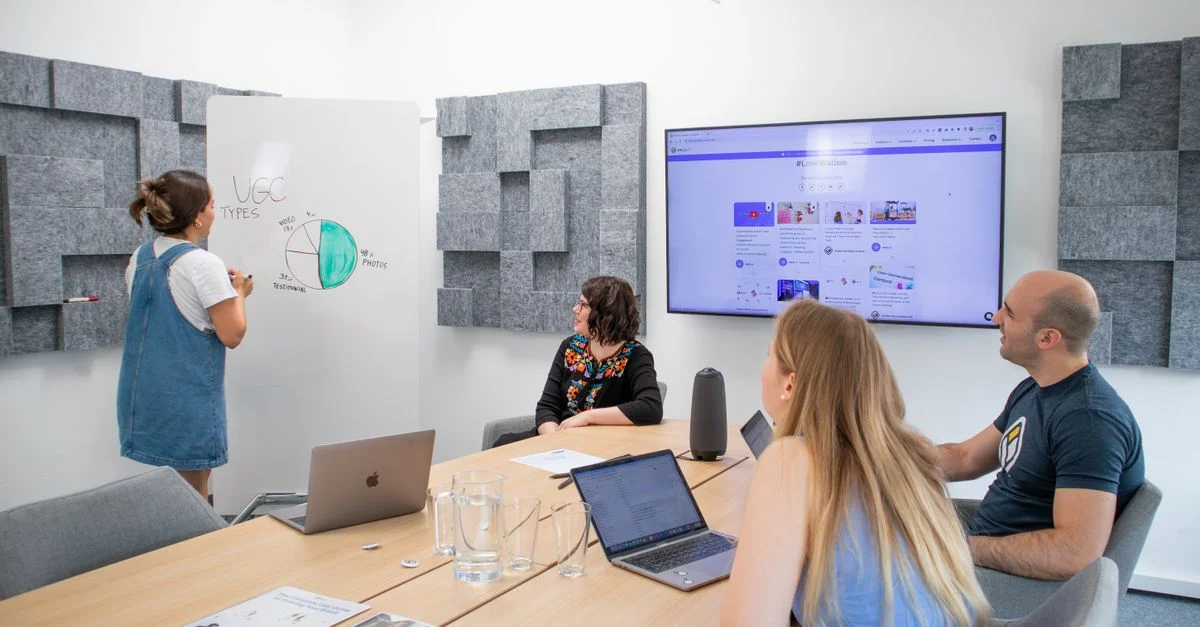 Des membres de l'équipe participant à une réunion moderne dans un bureau, discutant sur un tableau blanc et une présentation à l'écran.